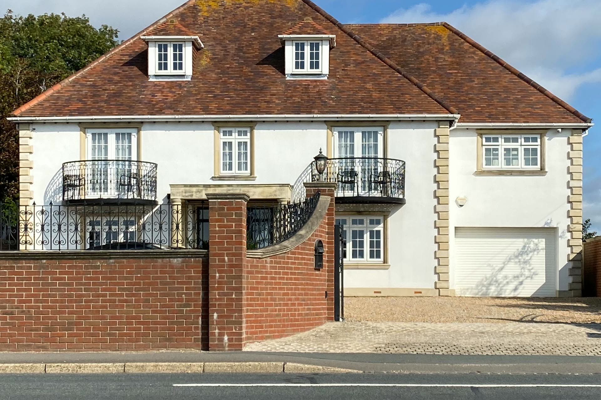 A substantial detached UK home with white render, stone quoins and a hipped tile roof, framed by a brick boundary wall